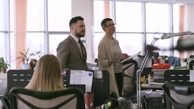 Two middle-aged businesspeople walking through modern office and discussing work, woman holding clipboard with documents