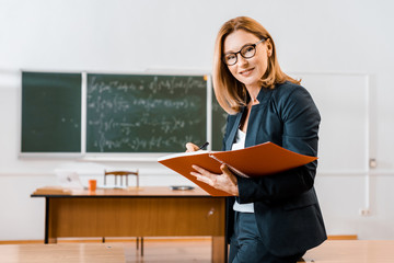 beautiful female teacher in formal wear writing in notebook and looking at camera in classroom