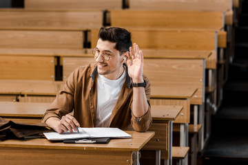 smiling male student in glasses sitting at desk and raising hand during lesson in classroom