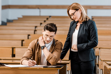 female university teacher looking at male student writing exam in classroom