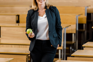 cropped view of female teacher in formal wear holding apple in classroom
