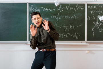 crumpled paper balls flying at frightened male teacher in classroom with chalkboard on background