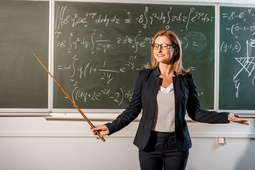 smiling female teacher with wooden pointer explaining mathematical equations in classroom