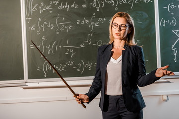 female teacher in formal wear with wooden pointer explaining mathematical equations in classroom