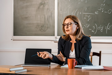 female teacher sitting at desk and pointing at laptop with blank screen in classroom