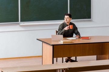 male teacher sitting at computer desk and drinking coffee in classroom