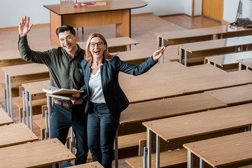 cheerful male student and female teacher with outstretched hands holding university books in classroom