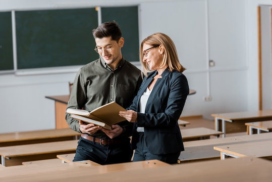 male student and female teacher in formal wear looking at university books in classroom
