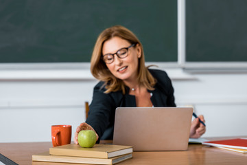 female teacher sitting at computer desk and reaching for apple in classroom