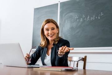 female teacher sitting at computer desk, holding glasses and looking at camera in classroom