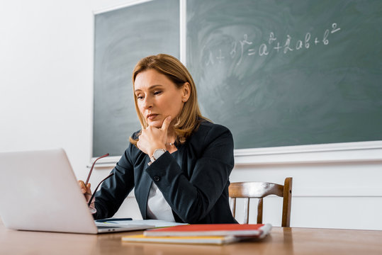 female teacher sitting at desk and using computer during lesson in classroom - Powered by Adobe