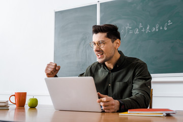 excited male teacher sitting at desk and using laptop in classroom