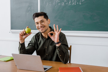 smiling male teacher in glasses holding apple and showing ok sign at computer desk in classroom