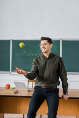 smiling male teacher throwing apple in class with chalkboard and desk on background