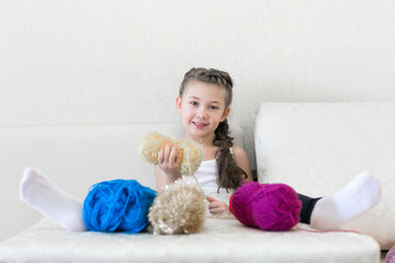 A girl with colored threads is sitting on the sofa. Beautiful little girl put her legs on the table and coils the thread.