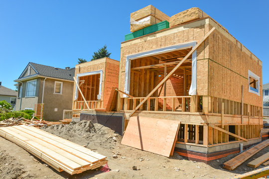 A single family home under construction. The house has been framed and covered in plywood. Stacks of board timber in front and stack of 2x4 boards on the top.