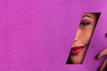 Girl with bright make up, red lipstick looking through hole in violet paper. Make up artist, beauty concept. Ready to party. Cosmetics sale. Beauty salon advertising banner with copy space - obrazy, fototapety, plakaty