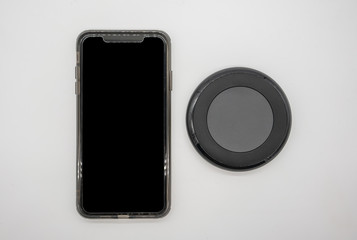 Black mobile phone placed by black round shape wireless charger isolated on white.