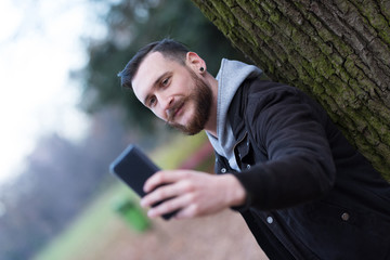 Fototapeta premium A handsome young hipster man leaning against a tree and taking a selfie in the park.