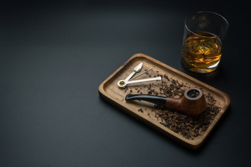 a smoking pipe with pipe tamper tool and tobacco in wooden tray, a glass of bourbon whiskey on the black table