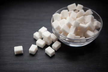 Fototapeta na wymiar White sugar cubes in little glass bowl on black textured background
