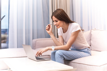 Fototapeta premium Happy female person is sitting on sofa and typing on laptop. She looking at screen with wide smile. Copy space on left side