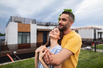 Joyful man is hugging his girlfriend and looking ahead with hint of smile on his face. Houses are on blurred background. Copy space on left side
