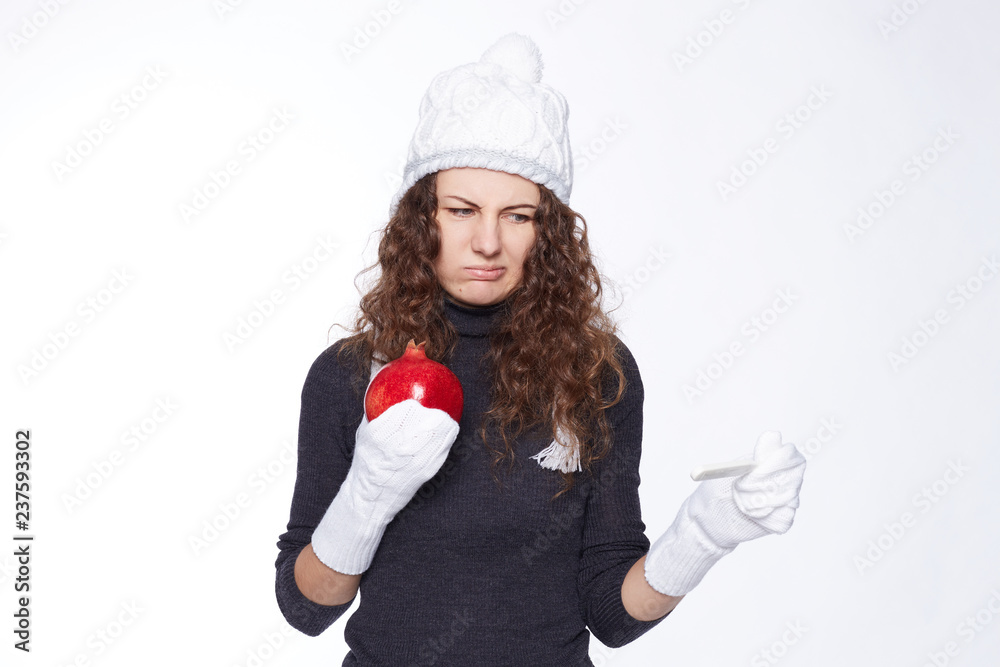 Wall mural Shocked female in winter outfit refuses to be ill with disgusting face expression. In full disbelief with negative grimace young woman looks at thermometer, holds pomegranate as medicine in hand.