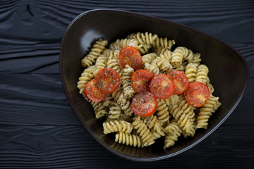 Bowl of italian fusilli served with basil pesto sauce and cherry tomatoes, flatlay on a black wooden background