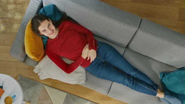 Girl in Red Jumper is Lying Down on a Sofa with Pillows. Looks Above and Smiles. Cozy Living Room with Modern Interior with Carpet, Plants, Coffee Table and Wooden Floor. Top View with Zoom In.