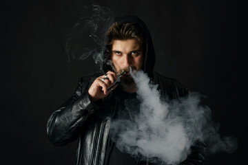 Man with vaping mod exhaling steam at black studio background. Bearded guy smoking e-cigarette to...