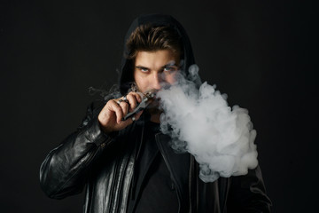 Man with vaping mod exhaling steam at black studio background. Bearded guy smoking e-cigarette to...