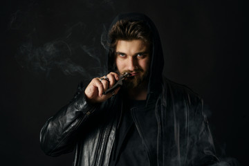Man with vaping mod exhaling steam at black studio background. Bearded guy smoking e-cigarette to quit tobacco. Vapor and alternative nicotine free smoking concept