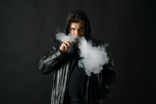 Man with vaping mod exhaling steam at black studio background. Bearded guy smoking e-cigarette to quit tobacco. Vapor and alternative nicotine free smoking concept