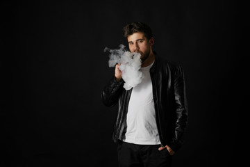 Man with vaping mod exhaling steam at black studio background. Bearded guy smoking e-cigarette to...