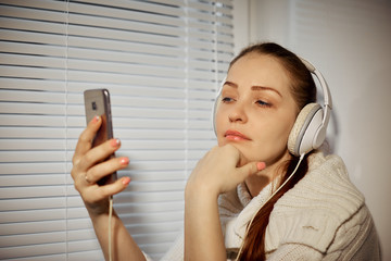 rest and relaxation, young Caucasian woman listening to music in headphones using a smartphone sitting on the windowsill