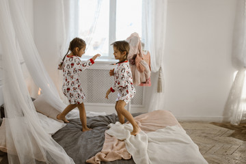 Two little girls in their pajamas are having fun jumping on a bed in a sunlit cozy bedroom