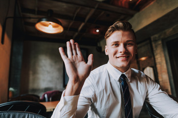 Concept of success and positive mind. Waist up portrait of young smiling businessman saying hi to somebody while sitting in cafe