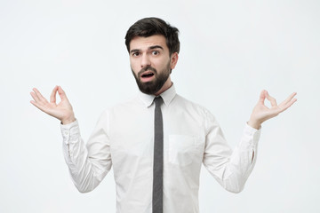 Studio portrait of young handsome male in white shirt, in meditation pose, trying to relax or calm down in zen, but he is feeling very annoyed. Yoga technic does not work.