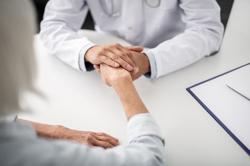 Fototapeta premium Close up of kind doctor sitting at the table in medical uniform and holding hand of his patient