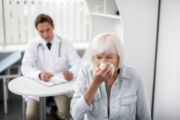 Obraz premium Elderly lady putting napkin to her nose and sneezing while the doctor sitting on the background
