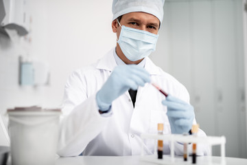 Serious doctor in medical mask looking and holding blood sample in test tube