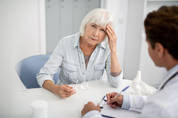 Tired elderly woman visiting her practitioner and touching the forehead while having headache