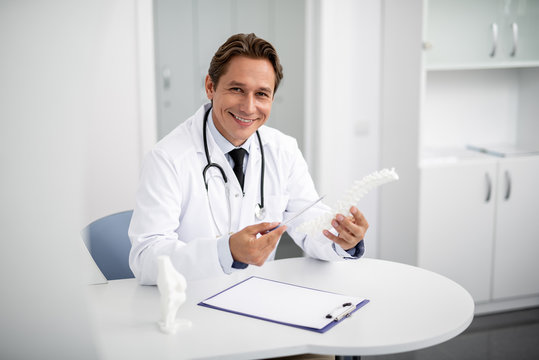 Cheerful general practitioner sitting at the table with spinal model in his hands and smiling while pointing to it
