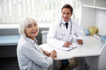 Obraz premium Positive emotional elderly lady turning her head and smiling while her practitioner sitting on the background
