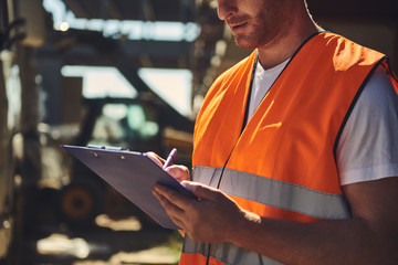 Close up of professional builder in orange uniform standing outdoors and carefully making notes while holding clipboard - Powered by Adobe
