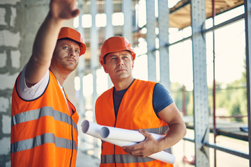 Attentive confident builder pointing to the distance while standing with his reliable coworker in unfinished building