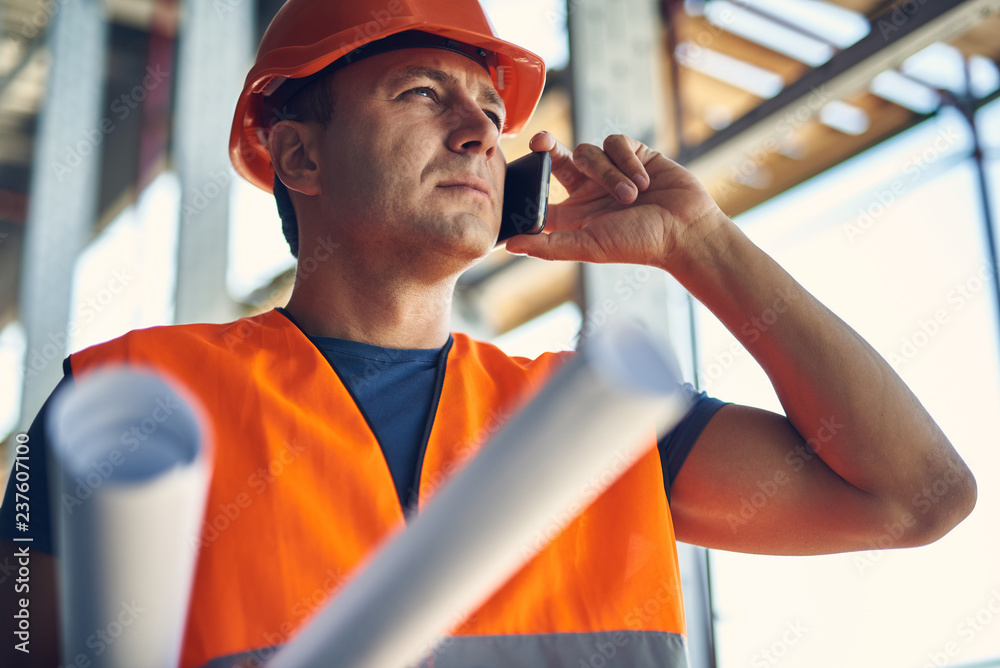 Wall mural Calm professional builder in orange helmet and vest putting his head up and frowning while talking on the phone