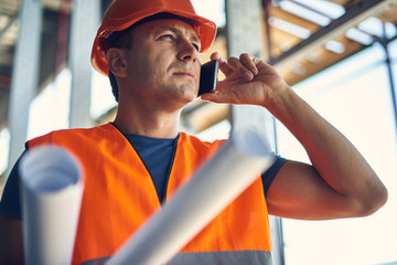Calm professional builder in orange helmet and vest putting his head up and frowning while talking...