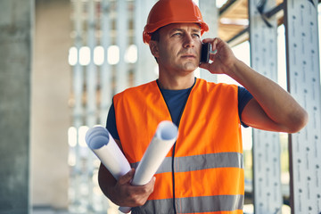 Phone talk. Thoughtful attentive builder in orange uniform standing with the drawings and having a...